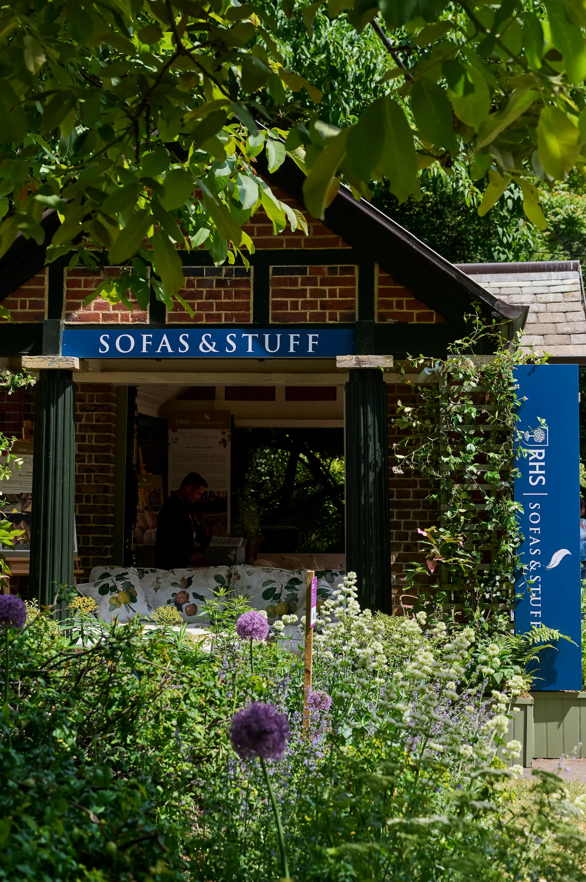 A brick garden pavilion with a blue sign reading Sofas & Stuff stands amid lush greenery and blooming flowers; a person is visible inside, partially obscured by a floral-print sofa.